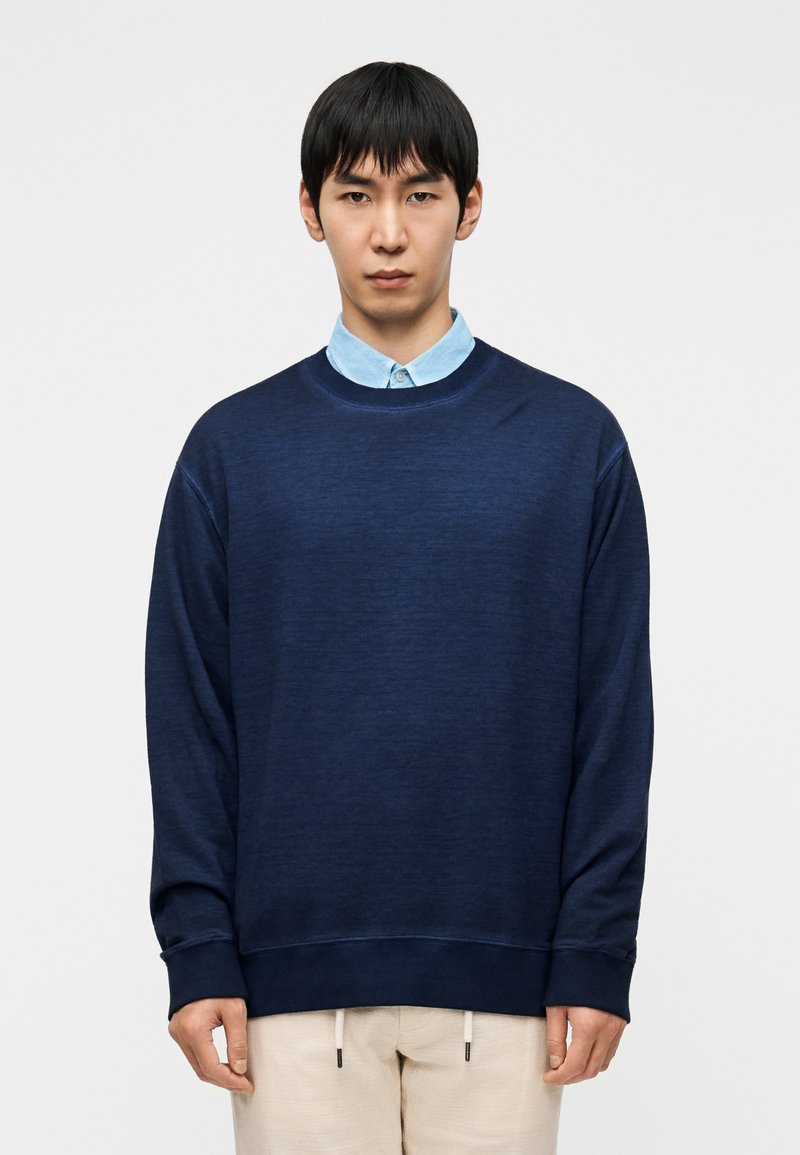 Μπλε σκούρο φούτερ με στρογγυλή λαιμόκοψη, μακρι ά μανίκια και υφασμάτινη υφή, φορεμένο πάνω από ένα ανοιχτό μπλε πουκάμισο με γιακά. Κανονική εφαρμογή.