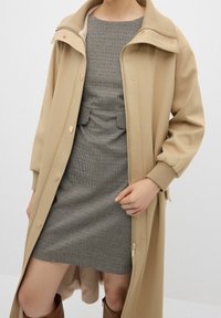 Abrigo exterior beige con cuello alto y botones dorados, superpuesto a un vestido ajustado de cuadros. Textura suave y diseño a medida.