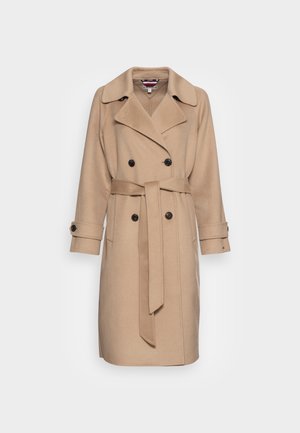 Beige dubbelgeknoopt lange jas met brede kraag, zwarte knopen, bijpassende riem en met knopen gesloten manchetten op een effen achtergrond.