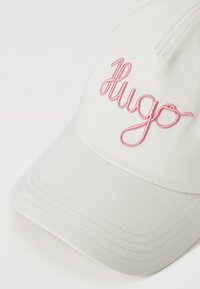 Valkoinen baseball-lippis, jossa on vaaleanpunainen brodeerattu "Hugo"-logo. Siinä on jämäkkä muoto ja ommeltu yksityiskohta lippahatussa.