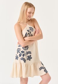 Ärmelloses beigefarbenes Kleid mit marineblauem Blumenmuster, mit einem Rüschensaum und einem Kreuzträgerdesign. Glatte Textur, lässige Silhouette.