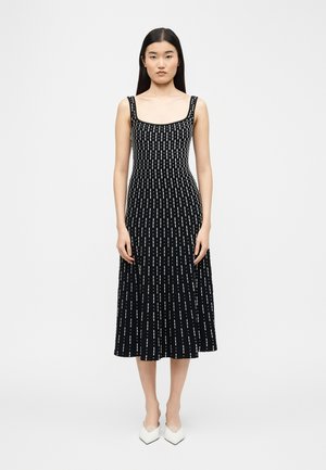 Mujer con cabello largo y negro, vestida con un vestido midi negro sin mangas con líneas blancas discontinuas verticales y zapatos blancos de punta puntiaguda, de pie mirando hacia adelante.