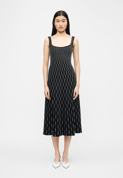 Femme aux longs cheveux noirs portant une robe midi noire sans manches avec des lignes blanches en pointillés verticales et des chaussures blanches à bouts pointus, debout face à l'avant.