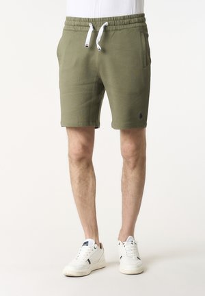 Uomo che indossa pantaloncini verde oliva con coulisse e sneakers bianche, in piedi contro uno sfondo chiaro e semplice.