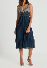 Námořnické plisované šaty s přiléhavým korzetem zdobeným stříbrnými a černými korálky, tenkými ramínky a sukní až do poloviny lýtek. Stříbrné sandály na podpatku.