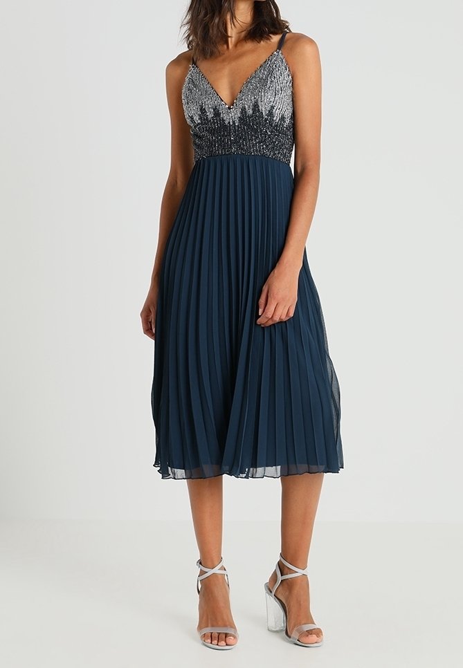 Námořnické plisované šaty s přiléhavým korzetem zdobeným stříbrnými a černými korálky, tenkými ramínky a sukní až do poloviny lýtek. Stříbrné sandály na podpatku.