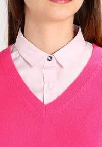 V-Ausschnitt Pulli aus leuchtend pinkem Strick, über einem hellpinken Hemd mit Kragen, das eine Knopfverschluss mit blauen und weißen Akzenten hat.