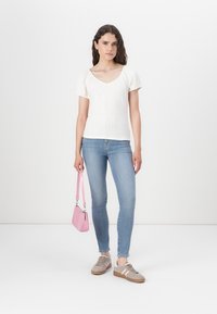 Bílá žebrovaná blůza s krátkými nafouknutými rukávy, kombinovaná se světle modrými skinny džíny a taupe teniskami. Drží malou růžovou kabelku.