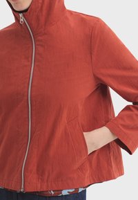 Veste rouge raccourcie avec une capuche, équipée d'une fermeture éclair argentée, de grandes poches avant et d'un tissu légèrement texturé.