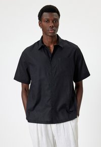 Camisa negra de manga corta con cuello abotonado, un bolsillo en el pecho y tela texturizada. El acabado suave realza el diseño ligero.