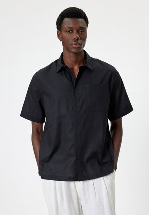 Chemise noire à manches courtes avec col boutonné, poche poitrine simple et tissu texturé. La finition lisse met en valeur le design léger.