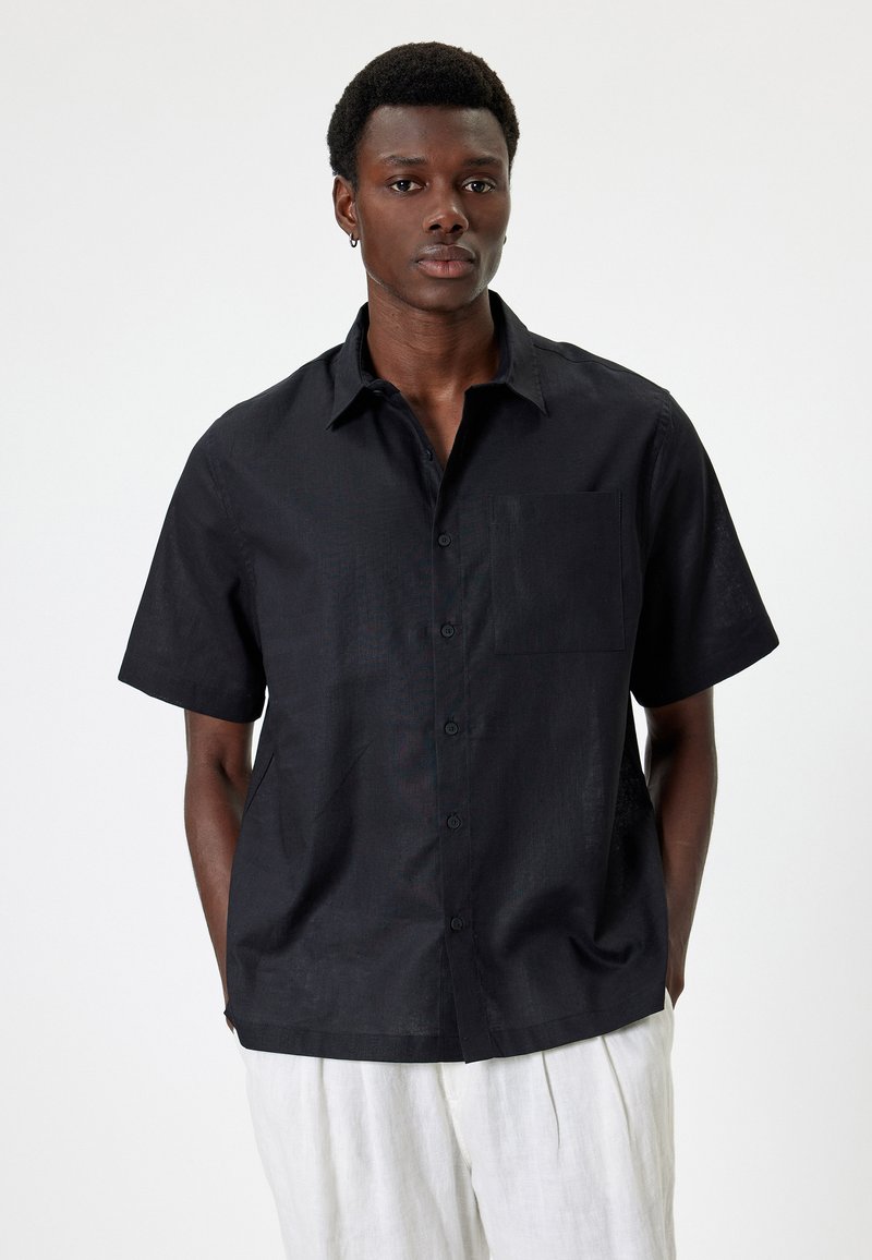 Chemise noire à manches courtes avec col boutonné, poche poitrine simple et tissu texturé. La finition lisse met en valeur le design léger.