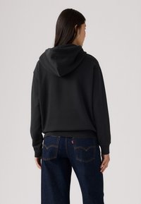 Sudadera negra con capucha ajustable. Tejido suave, corte holgado y puños de canalé. Combinada con unos vaqueros de pierna ancha azul oscuro que tienen bolsillos traseros.