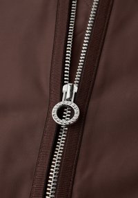 Nærbillede af en delvist lynet mørkebrun jakke med en sølvfarvet cirkulær lynlås trækkert, der har indgraveret tekst.