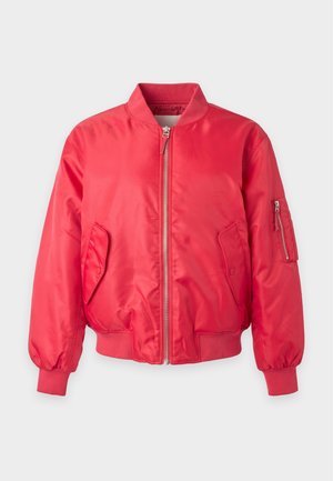 Femme aux longs cheveux bruns porte une veste bomber rouge, une chemise blanche, un jean bleu, des bottes noires et des boucles d’oreilles créoles, debout devant un fond uni.