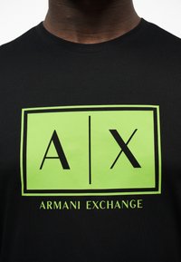 Μαύρο πουκάμισο με μεγάλο πράσινο ορθογώνιο λογότυπο που δείχνει τα γράμματα "A" και "X" διαχωρισμένα με μια κάθετη γραμμή, και την επιγραφή "ARMANI EXCHANGE" από κάτω.