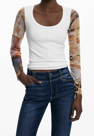 Personne portant un haut blanc côtelé à encolure dégagée avec des manches longues transparentes à fleurs multicolores et un jean en denim bleu foncé, main dans la poche.