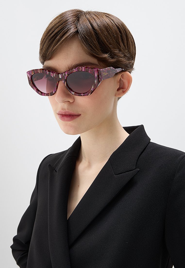 Fioletowe okulary przeciwsłoneczne w wzory z ciemnymi soczewkami, wyrazisty kształt kociego oka i teksturowane oprawki. W komplecie z czarnym żakietem.