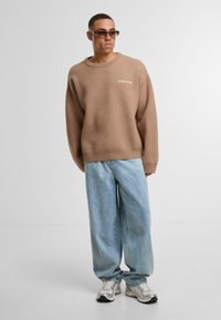 Hnědý oversize svetr s kulatým výstřihem, světle modré džínové kalhoty se širokými nohavicemi a béžové tenisky; svetr má logo "ANOTHER COTTON".