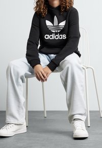 Černá mikina s bílým logem Adidas, spárovaná se světle modrými volnými džíny a bílými teniskami. Model sedí na kovové židli.