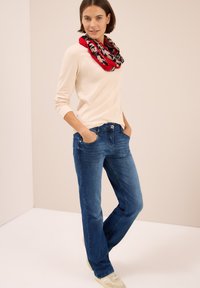 Haut beige à manches longues, jeans bleus et écharpe à motifs multicolores. Les jeans ont une coupe droite et un design à cinq poches.