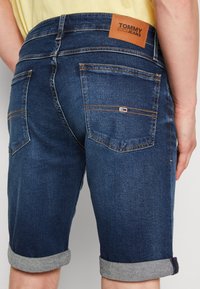 Person wearing rolled-up dark blue denim shorts with Tommy Jeans leather patch and back pockets visible, standing against a plain background.