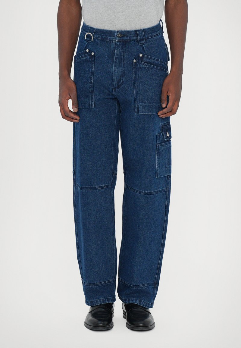 Pantalones cargo de denim azul oscuro con múltiples bolsillos, corte holgado y herrajes resistentes. Presentan un cierre de botón y detalles de costura visibles.