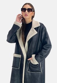 Abrigo de cuero negro con un cuello y bordes de bolsillo de borrego sintético crema. Largo, de corte relajado y sin botones en el frente.