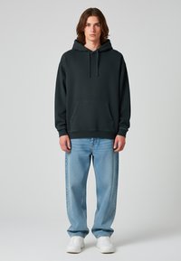 Felpa grigia scura con tasca a marsupio e coulisse regolabile, abbinata a jeans chiari a vestibilità ampia e sneakers bianche.