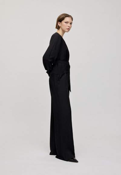 Zwarte wikkel-top met lange mouwen en zwarte wijde broek. Glad materiaal, minimale uitstraling en zwarte pumps met puntige neus.