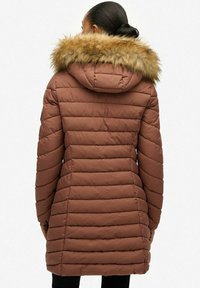 Brązowy puchowy płaszcz z kapturem, z futrzanym wykończeniem. Projekt ma przeszywane poziome panele i średnią długość.