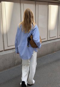 Blue and white striped oversized shirt, cream wide-leg pants, holding a brown leather bag, walking on a textured pavement.