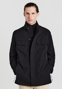 Giacca leggera nera con due tasche sul petto, colletto alto e zip frontale. Presenta una superficie liscia e cordini regolabili all'orlo.