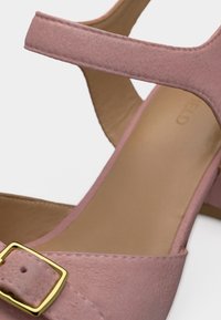 Κοντινό πλάνο ενός ροζ σουέντ σανδαλιού με χρυσή αγκράφα και μαξιλαρωτό μπεζ πάτο, που διαθέτει λουράκι γύρω από τον αστράγαλο.