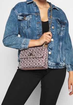 Kobieta w niebieskiej dżinsowej kurtce i czarnym stroju, trzyma beżową torebkę Calvin Klein z monogramem i złotym łańcuszkiem.