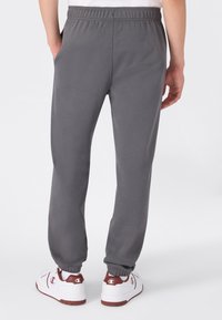 Persoon van achter in grijze joggingbroek met elastische boorden en witte sneakers met bruine accenten, staande met ontspannen handen.