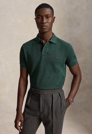 Jeune homme portant un polo vert foncé et un pantalon gris plissé à taille haute avec une montre au poignet gauche.