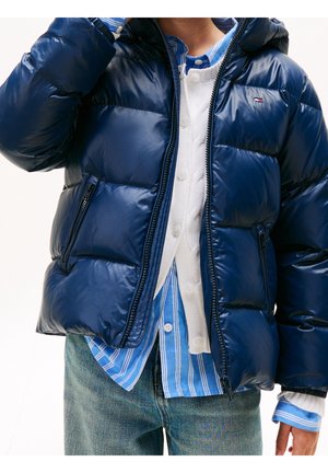 Personne portant une doudoune bleue marine brillante, un cardigan blanc boutonné, une chemise rayée bleue et un jean, avec un bras levé près du visage.