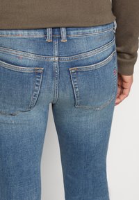 Jeans de denim azul con una cintura de altura media, costuras en contraste y dos bolsillos traseros. La tela tiene un aspecto desgastado con ligeros detalles de arrugas.