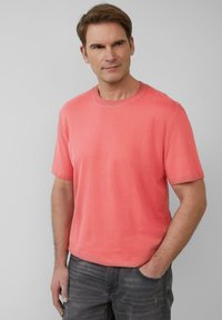 Ein Mann trägt ein korallenfarbendes T-Shirt mit kurzen Ärmeln und dunkle Jeans. Er steht mit einer Hand in der Tasche und schaut leicht zur Seite vor einem schlichten Hintergrund.