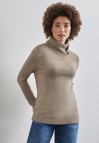 Beige rolák s žebrovaným límcem a lemem, vyrobený z měkké a strukturované tkaniny. Modelka má na sobě modré džíny.