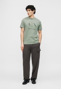 Νέος άνδρας που φοράει πράσινο φασκόμηλο T-shirt Armani Exchange και σκούρο γκρι παντελόνι καρότσα, στέκεται με το ένα χέρι στην τσέπη, κοιτάζοντας προς τα δεξιά.