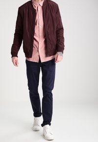 Burgundy bomberjacka över en ljusrosa knappskjorta, kombinerad med mörka byxor och vita sneakers. Mjukt tyg, avslappnad design.