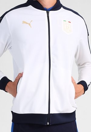 Weisse Sportjacke mit Reißverschluss und schwarzem Besatz, goldenes Puma-Logo auf der rechten Brust sowie ein Italien-Wappen auf der linken Brust, getragen mit den Händen in den Taschen.