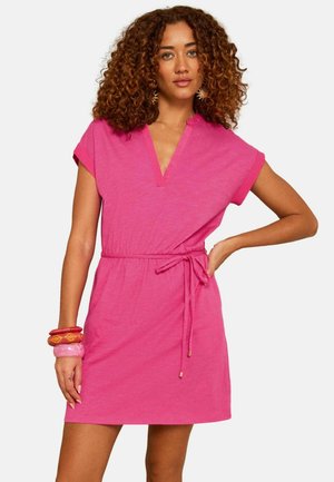 Vrouw met krullend haar die een korte mouwen roze jurk draagt met een taillekoord, gecombineerd met rode en roze armbanden en oorbellen in de vorm van de zon.