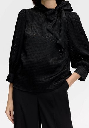 Zwarte blouse met een gestructureerd patroon, voorzien van een hoge kraag en een gestrikte strik bij de hals. Lange mouwen met geplooide manchetten.