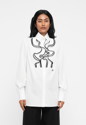 Camicia bianca con dettagli arricciati neri, maniche lunghe e colletto classico. Presenta un piccolo logo sul lato sinistro.