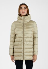 Giovane donna che indossa una giacca imbottita beige con colletto alto, in piedi su uno sfondo bianco semplice, rivolta in avanti con un'espressione neutra.
