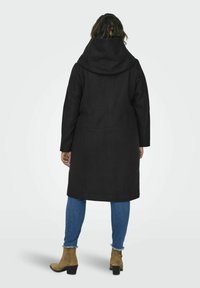 Manteau à capuche noir avec une forme structurée, des manches longues et une texture lisse, associé à un jean bleu et des bottines marron.