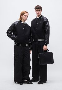 Jachete bomber negre cu mâneci din piele, manșete albe cu dungi, asortate cu pantaloni cargo negri și o servietă din piele, pe un fundal alb.